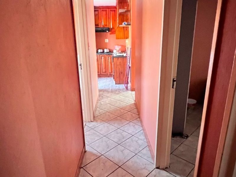A narrow hallway with tiled flooring leading towards a kitchen area featuring wooden cabinetry and an extractor fan.