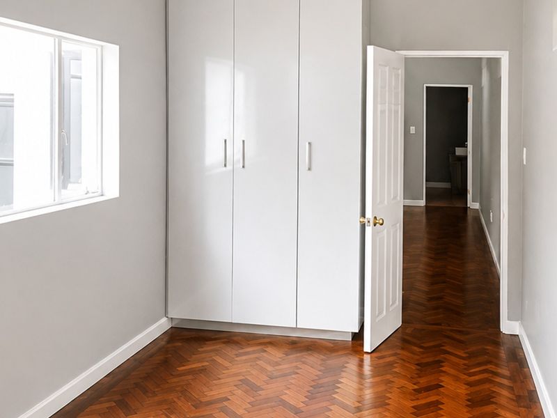 A bright, empty bedroom featuring polished herringbone parquet flooring, neutral walls, and modern built-in cupboards, with a view into the hallway.