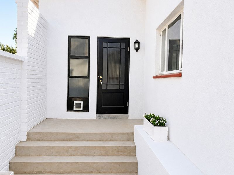 A clean and well-maintained cottage entrance featuring concrete steps, a black panelled front door with a side light, and exterior wall lighting.