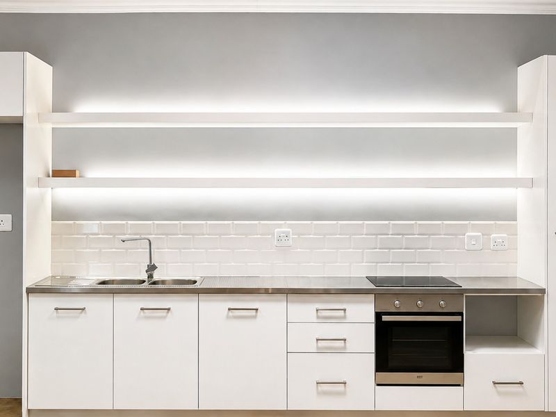 Modern kitchen featuring white cabinetry, stainless steel countertops, subway tile backsplash, built-in oven, hob, and integrated LED lighting on floating shelves.