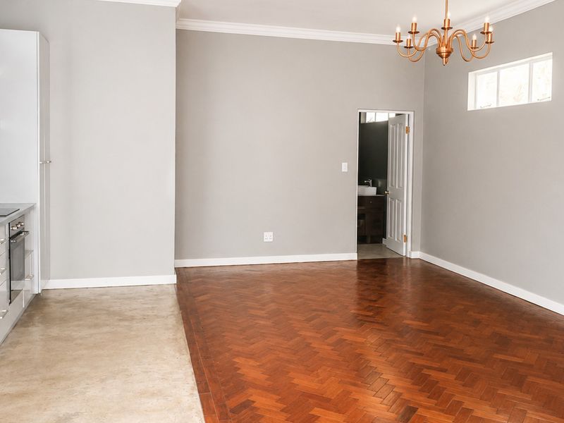 An open-plan living and kitchen area featuring polished herringbone parquet flooring, modern white cabinetry, and a classic chandelier, offering a clean, versatile space.