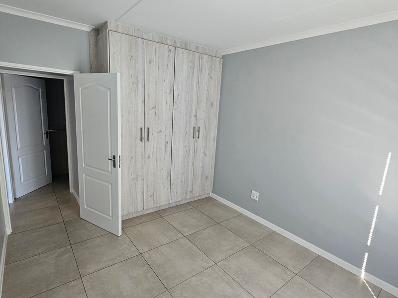 A clean, empty bedroom featuring neutral grey walls, large-format floor tiles, and modern wood-look built-in cupboards.