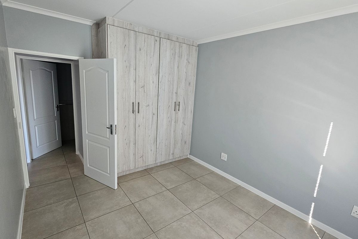 A clean, empty bedroom featuring neutral grey walls, large-format floor tiles, and modern wood-look built-in cupboards.