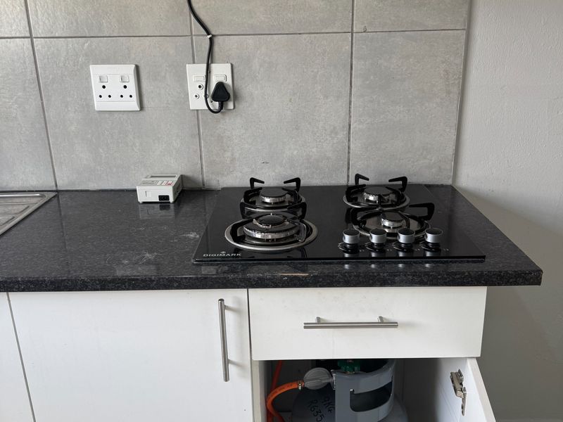 A functional kitchen area featuring a modern black gas hob, extractor fan, granite countertops, and tiled backsplash with integrated gas cylinder storage.