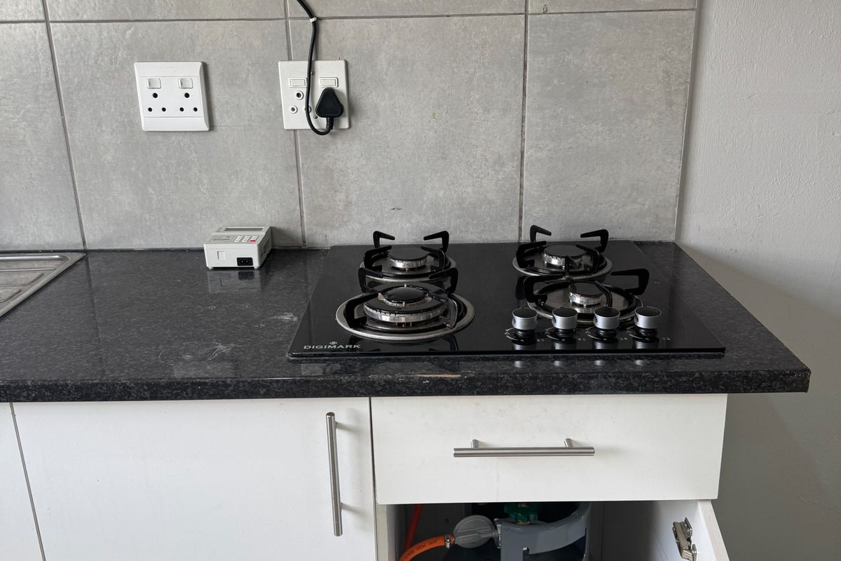 A functional kitchen area featuring a modern black gas hob, extractor fan, granite countertops, and tiled backsplash with integrated gas cylinder storage.