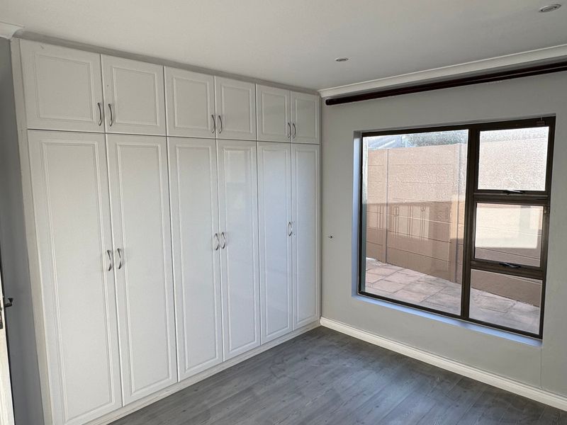 A clean, empty bedroom featuring modern laminate flooring, extensive white built-in cupboards, and a large aluminium-framed window providing natural light.