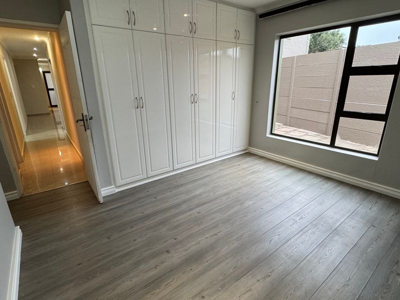 A bright, empty bedroom featuring modern laminate flooring, built-in white cupboards, and a large window with views of the exterior boundary wall.