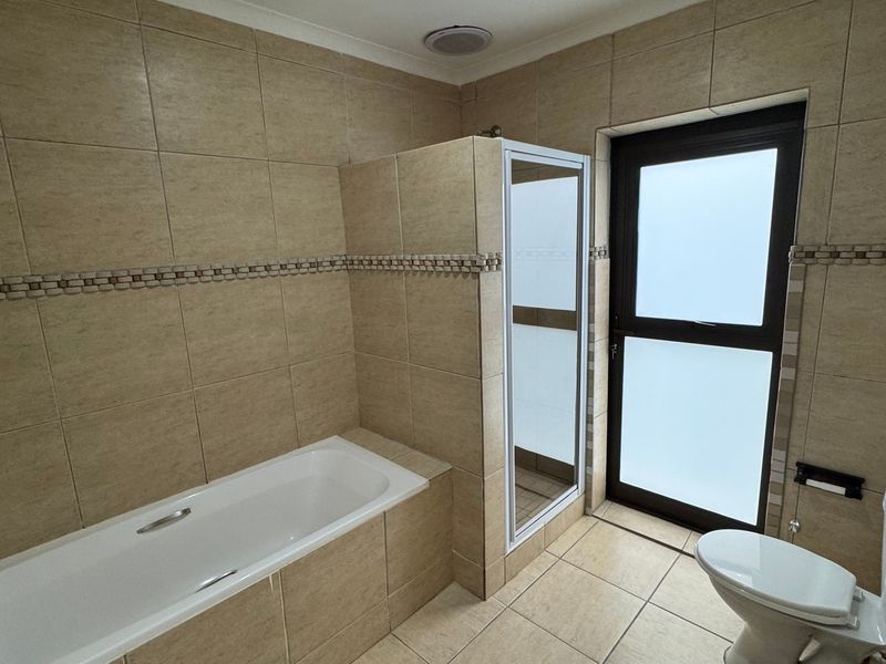 A clean, tiled bathroom featuring a built-in bathtub, a separate shower enclosure, and a toilet, with natural light provided by a frosted glass door.