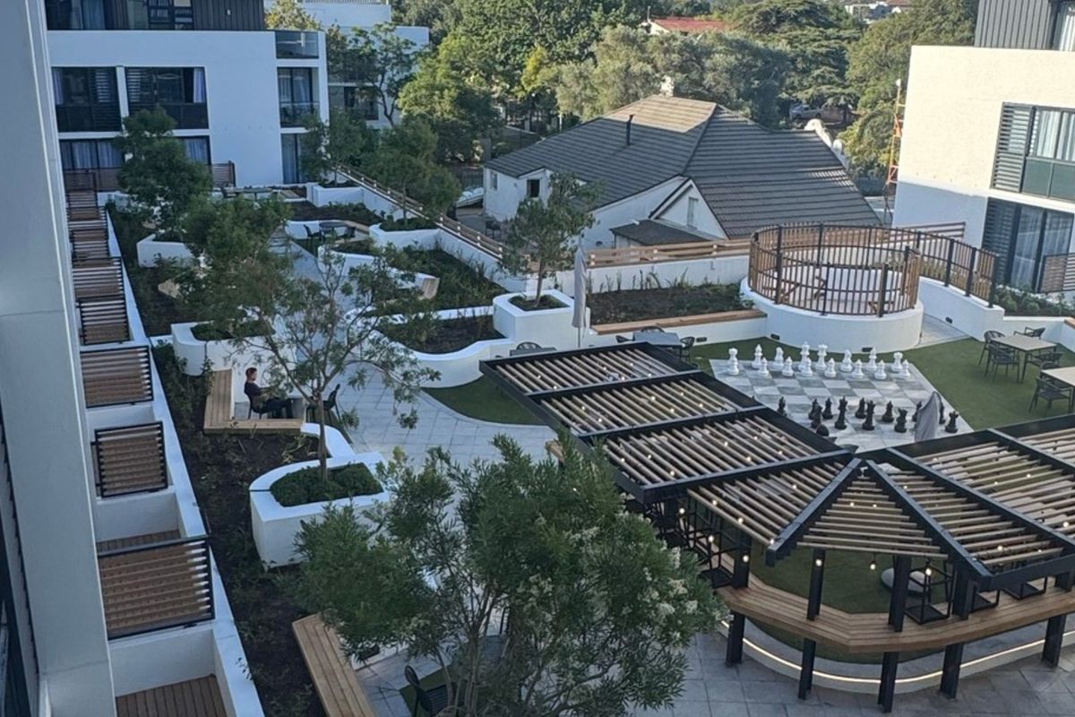 A well-maintained communal outdoor courtyard featuring landscaped gardens, paved pathways, a timber pergola, and a large outdoor chess set.