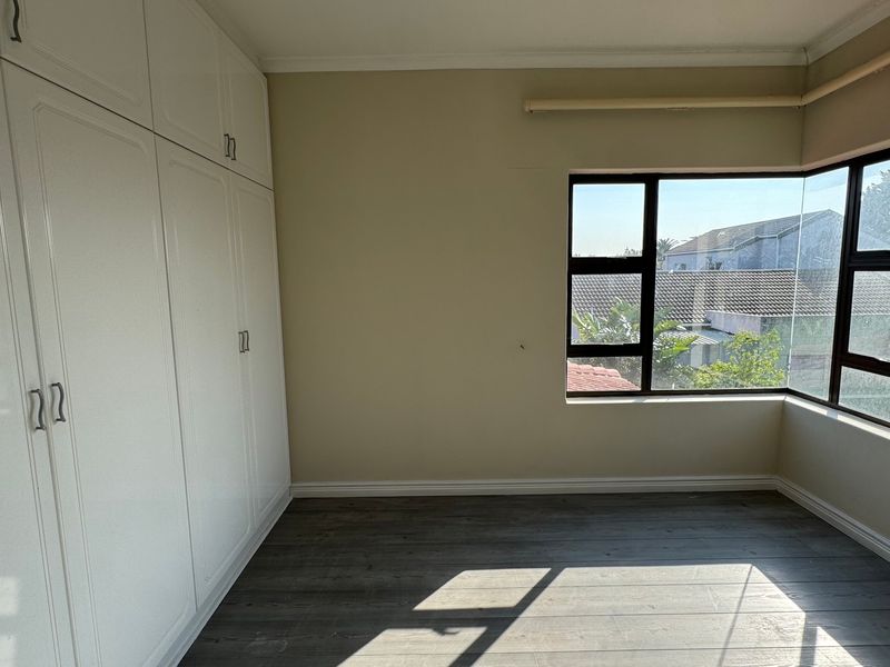 A bright, empty bedroom featuring modern laminate flooring, neutral walls, and extensive built-in cupboard storage with large windows.