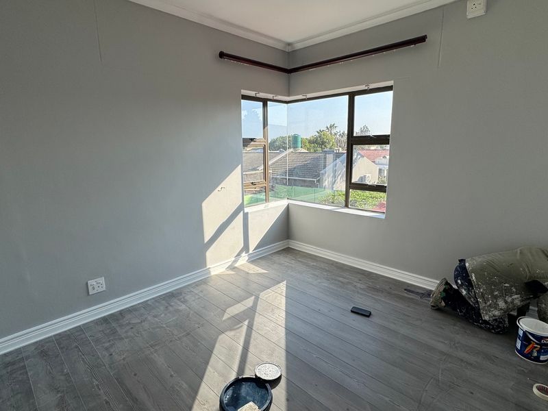A bright, empty bedroom featuring modern grey laminate flooring, neutral walls, and a large corner window providing natural light.