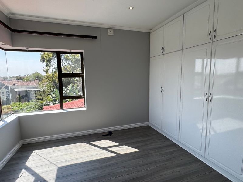 A bright and spacious bedroom featuring modern laminate flooring, extensive built-in white cabinetry, and a large bay window providing natural light.