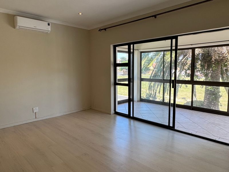 Bright and spacious living area featuring light laminate flooring, a wall-mounted air conditioner, and large sliding glass doors opening onto a covered patio.