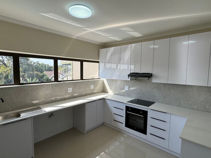 A modern, bright kitchen featuring sleek white cabinetry, stone countertops, integrated appliances, and a large window providing ample natural light.