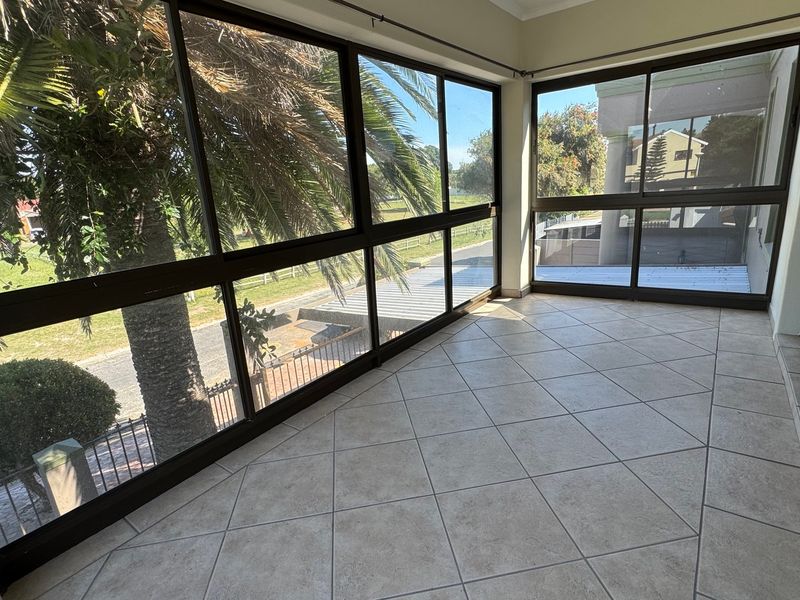 A bright, enclosed patio or sunroom featuring large glass windows, aluminum frames, and neutral tiled flooring, offering an airy space with garden views.