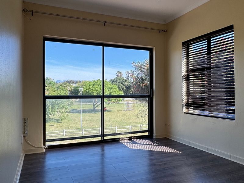 A bright, empty bedroom featuring dark laminate flooring, a large sliding glass door with garden views, and fitted venetian blinds.