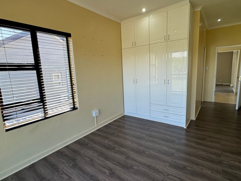 A bright, empty bedroom featuring modern white built-in cupboards, dark laminate flooring, and recessed downlighting, offering a clean canvas for personalization.
