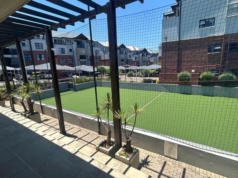 A well-maintained outdoor mini soccer pitch featuring artificial turf, enclosed by perimeter netting and a timber pergola structure within the apartment complex.