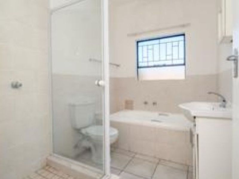 A functional bathroom featuring a shower enclosure, bathtub, vanity basin, and toilet, with neutral wall tiles and a window with security bars.