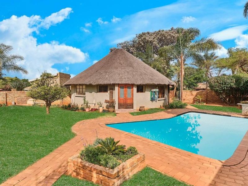 Rear exterior view featuring a thatched roof structure, a swimming pool with brick paving, and a landscaped garden area.