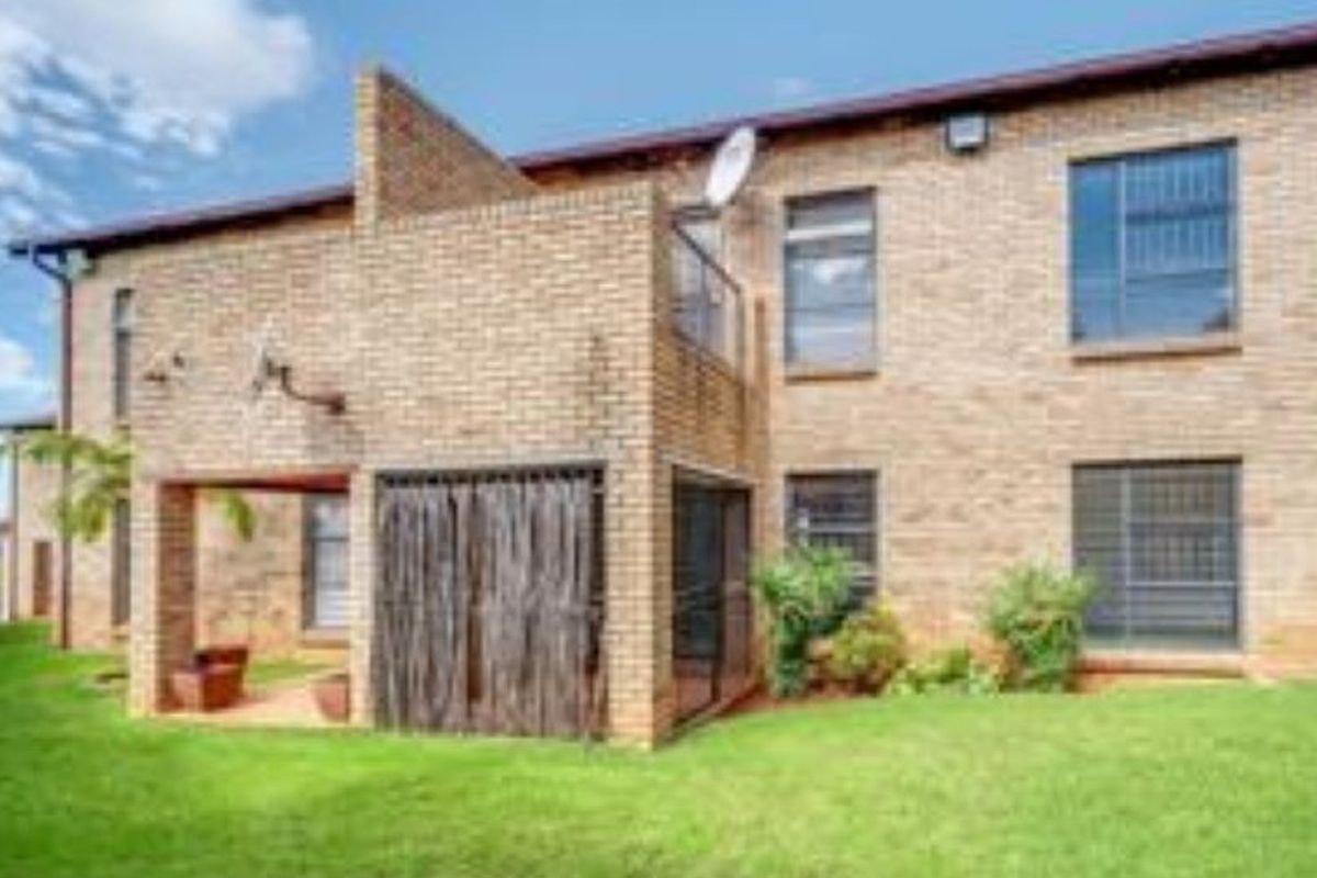Rear view of a face brick townhouse featuring a covered patio area, security bars on windows, and a well-maintained lawn.