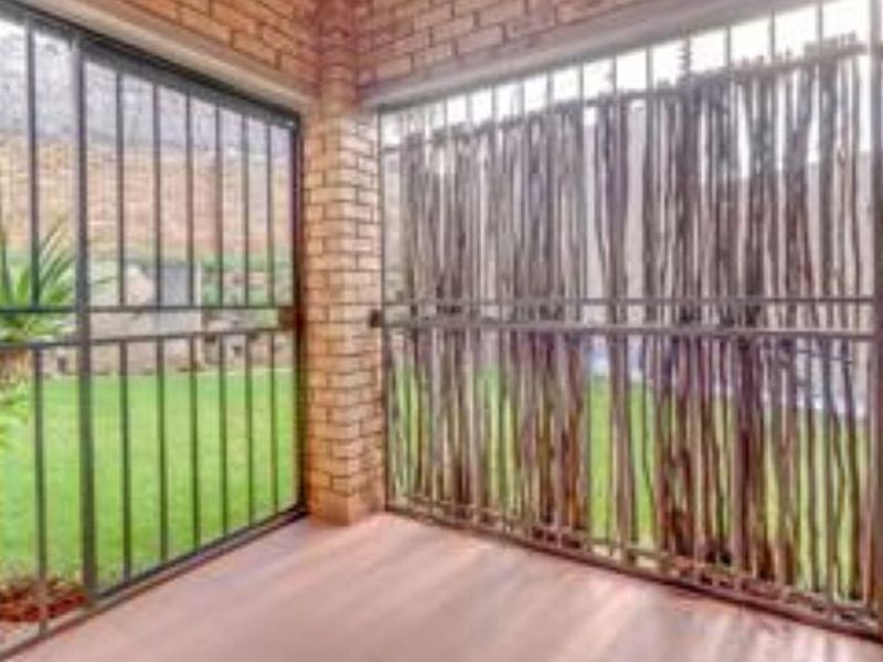 A covered patio area featuring exposed brick walls, laminate flooring, and security bars overlooking a private garden space.