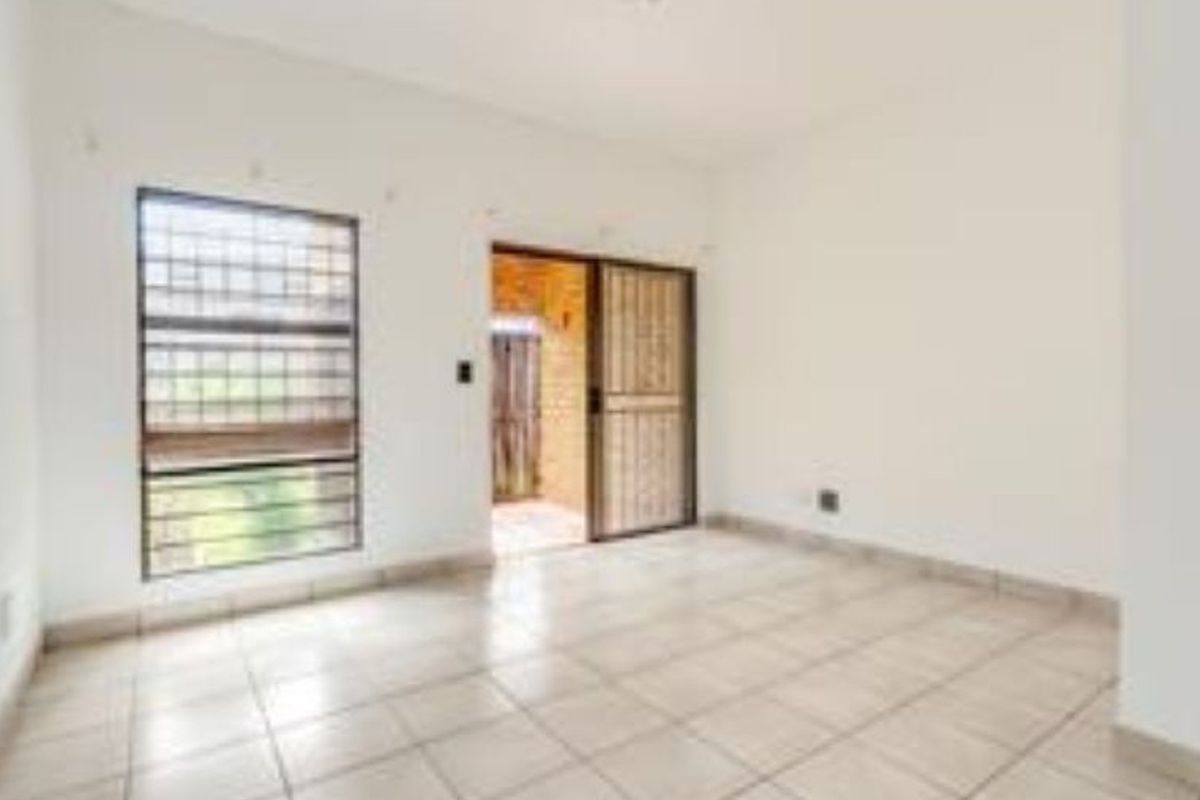 A bright, empty living room featuring tiled flooring, security bars on the window, and a sliding security gate leading to an outdoor area.