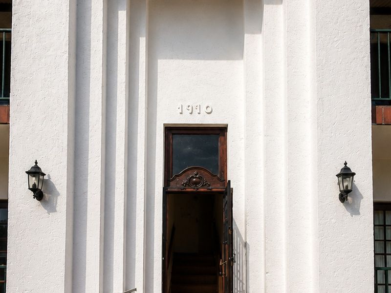 Front facade of a historic 1910 building featuring a decorative wooden entrance door, symmetrical balconies with wrought iron railings, and classic exterior wall lighting.