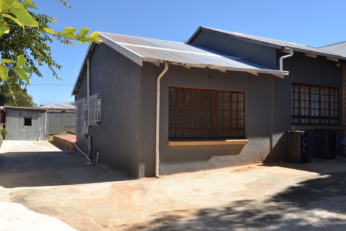 Rear exterior view of the cottage featuring a corrugated metal roof, timber-framed windows with security bars, and a concrete driveway area.