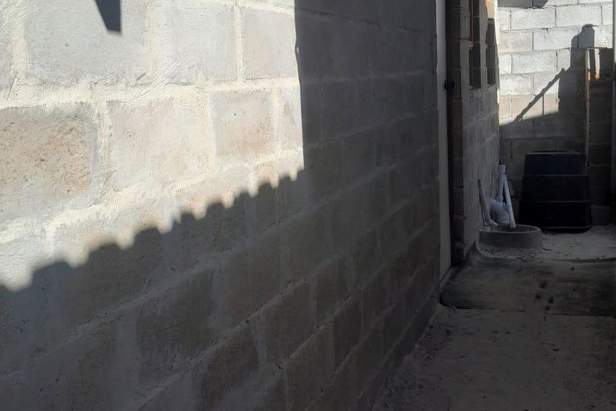 A narrow exterior side passage featuring unfinished concrete block walls and a concrete pathway with exposed plumbing infrastructure.