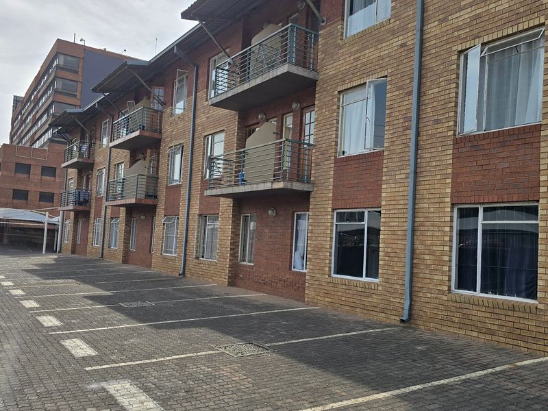 Exterior view of a multi-story brick apartment building featuring private balconies with metal railings and an adjacent paved parking area.
