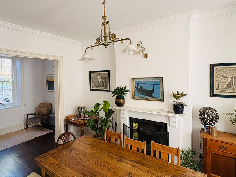 A well-maintained dining room featuring a classic fireplace with mantelpiece, ornate ceiling cornice, and polished hardwood flooring, leading into an adjacent living space.