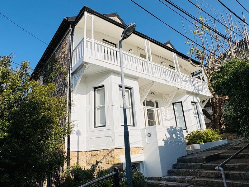 A charming two-story house featuring a white facade, a prominent upper-level balcony with timber balustrades, and a stone staircase leading to the entrance.