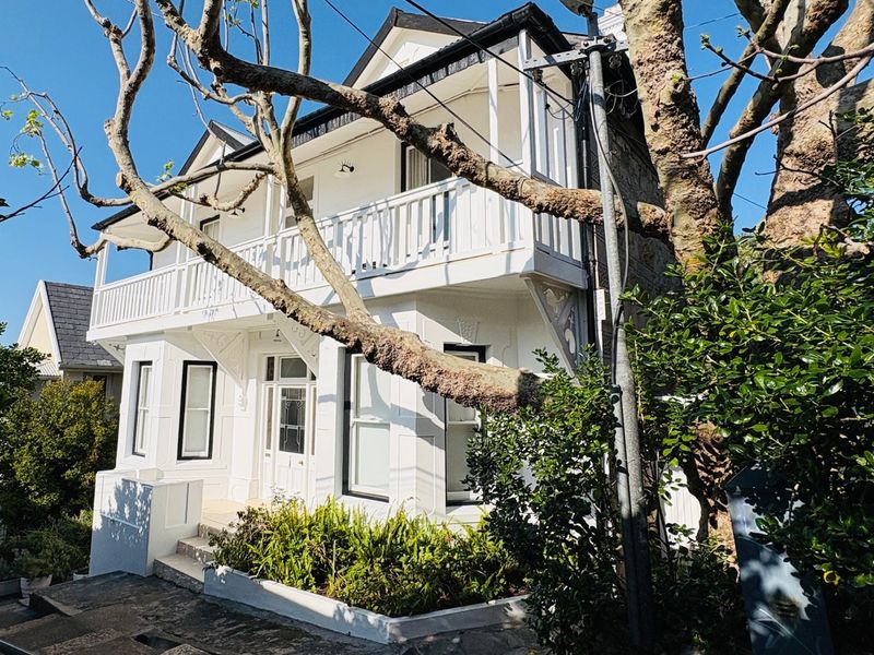 A charming two-story white house featuring a classic gabled roof, a prominent upper-level balcony with timber balustrades, and established garden landscaping.