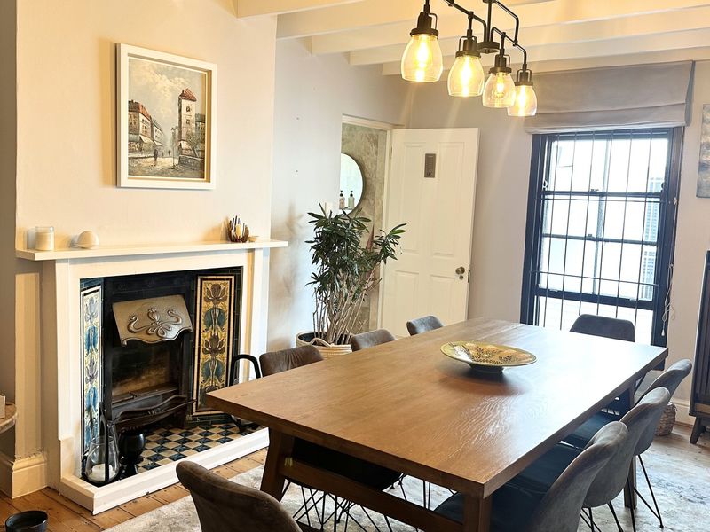 A charming dining room featuring a decorative fireplace with tiled hearth, exposed ceiling beams, and hardwood flooring, illuminated by a modern pendant light fixture.