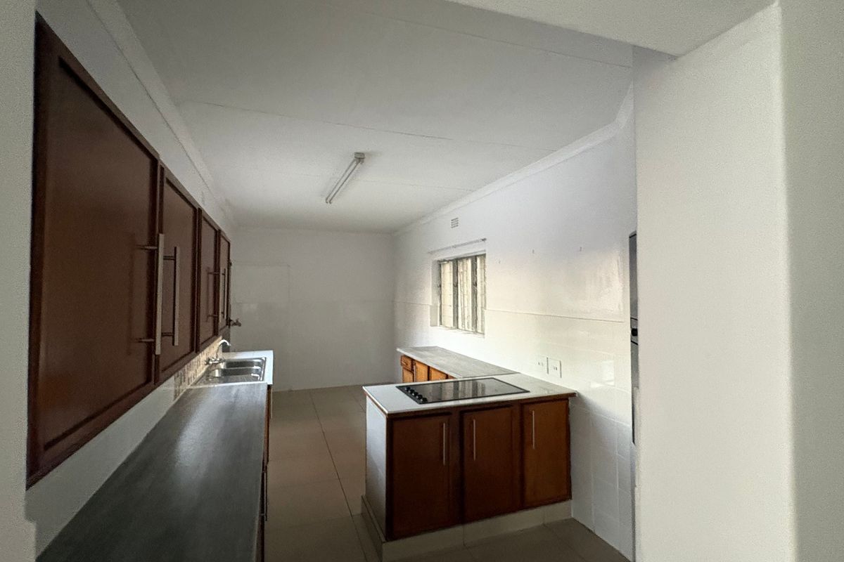 A functional, empty kitchen featuring built-in wooden cabinetry, a double sink, and a built-in hob with tiled splashbacks and flooring.