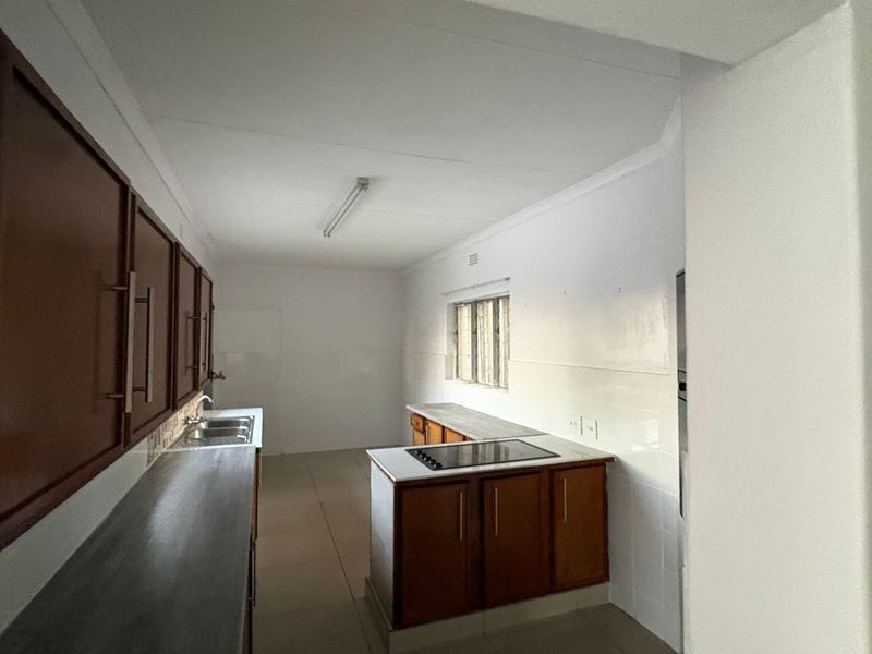 A functional, empty kitchen featuring built-in wooden cabinetry, a double sink, and a built-in hob with tiled splashbacks and flooring.
