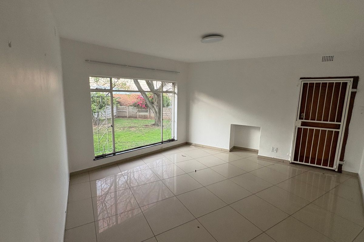 A bright, empty living room featuring neutral tiled flooring, a large window overlooking the garden, and a secure entrance door.