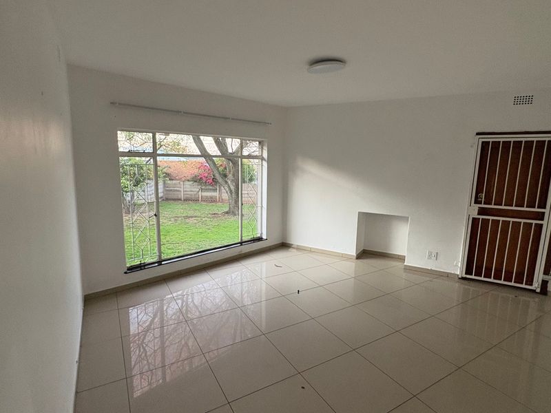 A bright, empty living room featuring neutral tiled flooring, a large window overlooking the garden, and a secure entrance door.