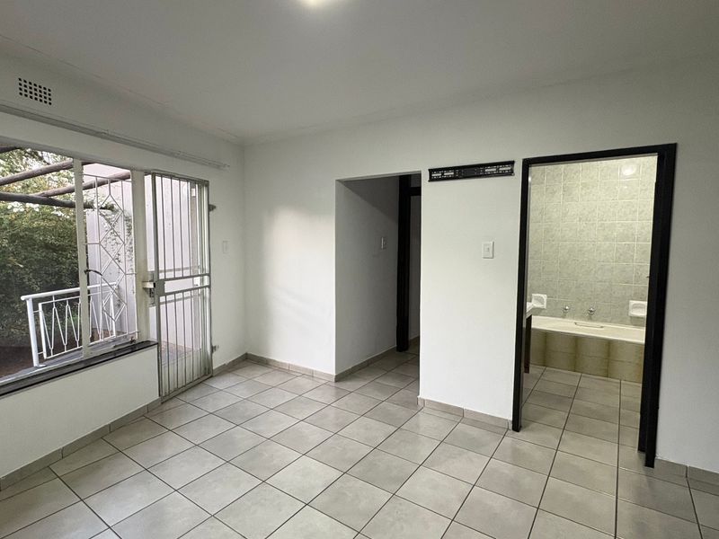 A bright, empty bedroom featuring tiled flooring, a large window with security bars, and direct access to an en-suite bathroom.
