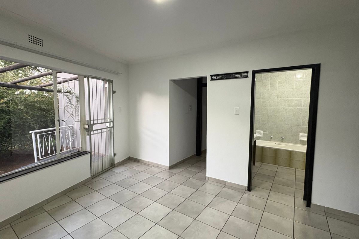A bright, empty bedroom featuring tiled flooring, a large window with security bars, and direct access to an en-suite bathroom.