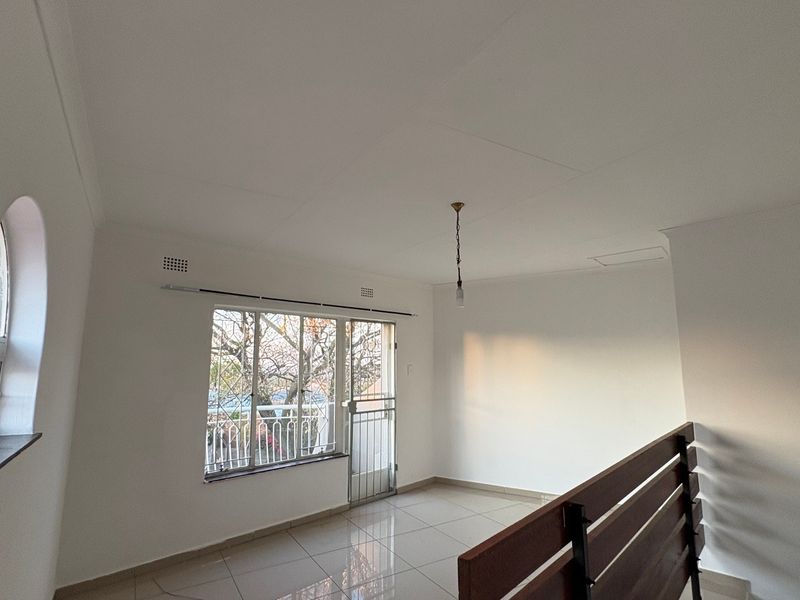 A bright, empty living area featuring tiled flooring, an arched window, and a wooden staircase railing, offering a clean and neutral space.