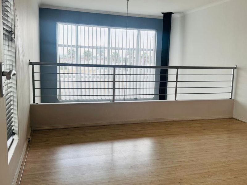 An empty living area featuring laminate flooring, a metal balustrade overlooking the space below, and large windows with vertical blinds.