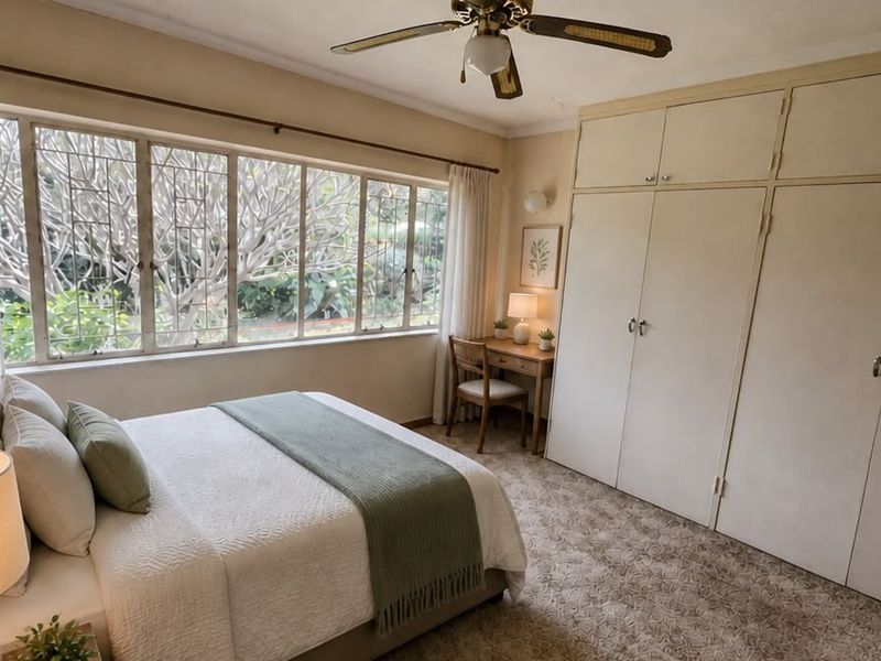 A well-lit bedroom featuring extensive built-in cupboards, a ceiling fan, and large windows overlooking the garden.