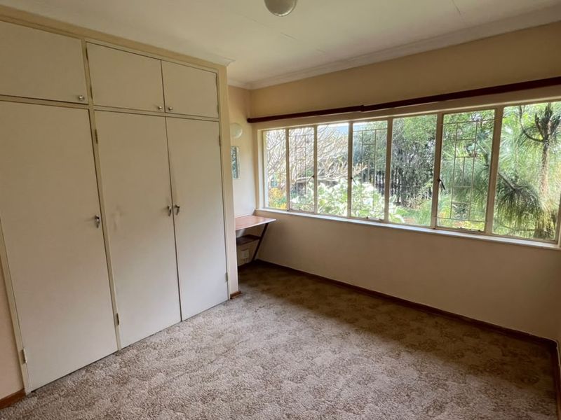 A bright, empty bedroom featuring built-in cupboards, carpeted flooring, and a large window with security bars overlooking the garden.