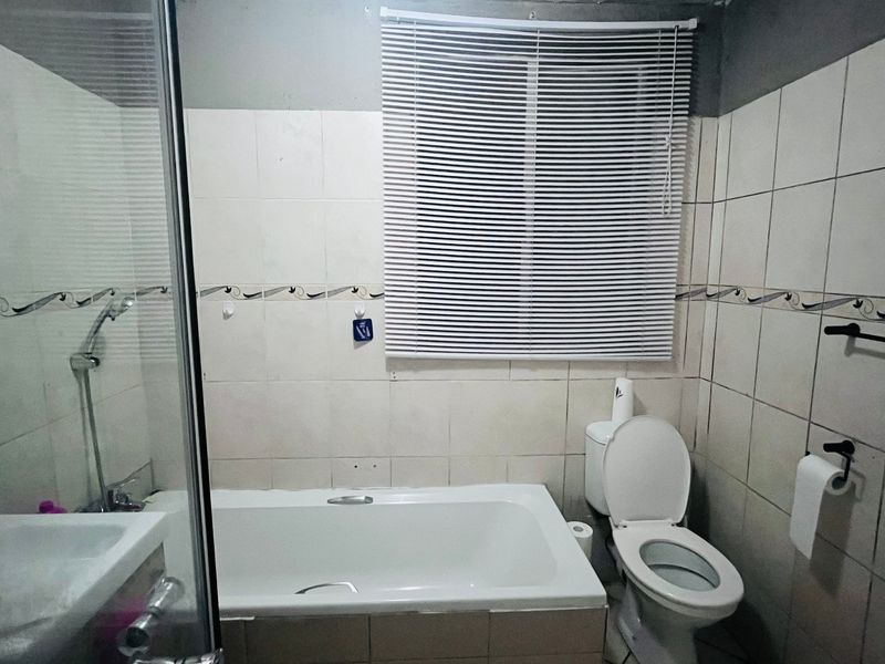A functional bathroom featuring a bathtub, shower enclosure, and toilet, with tiled walls and venetian blinds. The space would benefit from cosmetic updates.