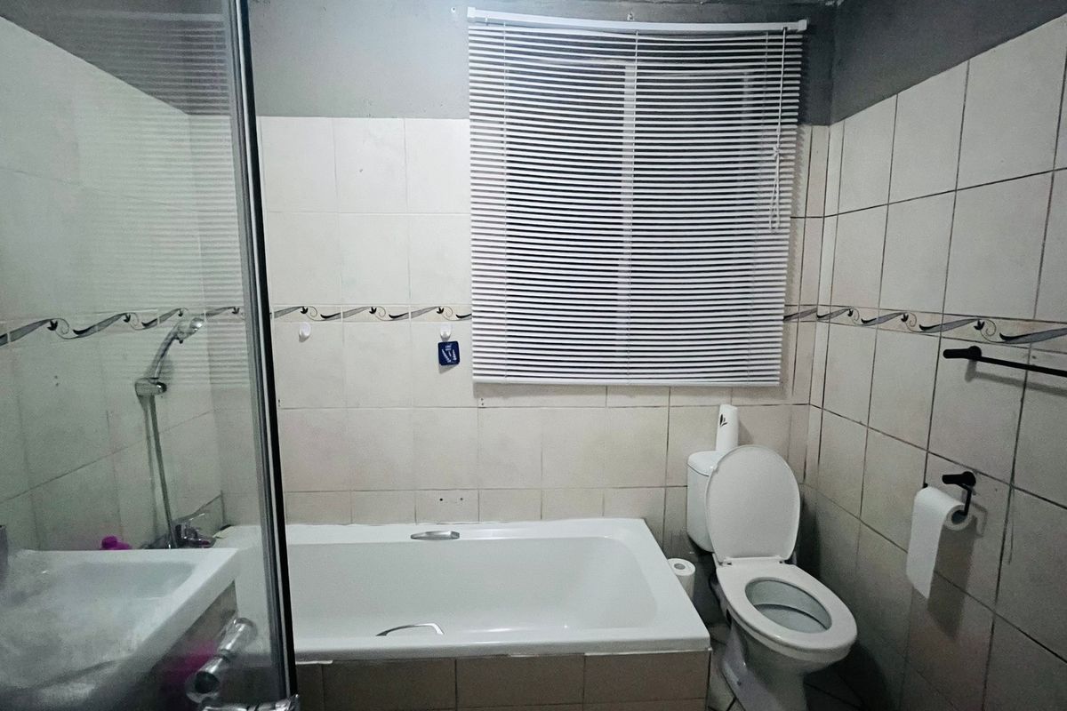 A functional bathroom featuring a bathtub, shower enclosure, and toilet, with tiled walls and venetian blinds. The space would benefit from cosmetic updates.