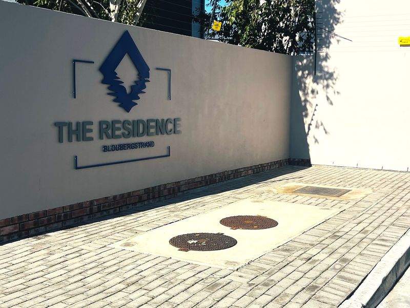 The entrance area of The Residence in Bloubergstrand, featuring a prominent boundary wall with signage, electric fencing, and a paved driveway.