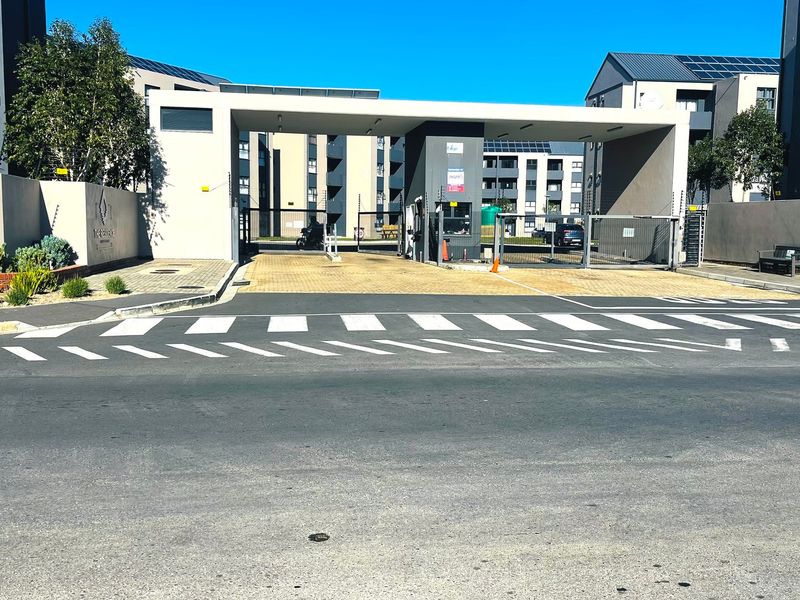 The main entrance and security gatehouse of the apartment complex, featuring paved access, perimeter walling, and modern architectural design.
