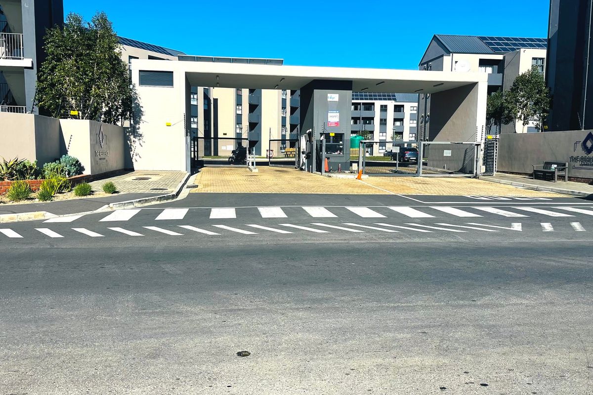 The main entrance and security gatehouse of the apartment complex, featuring paved access, perimeter walling, and modern architectural design.